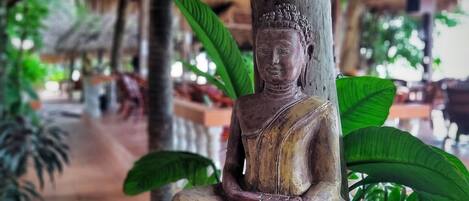 Una estatua de Buda en actitud meditativa, rodeada de follaje y una estructura con techo de paja.