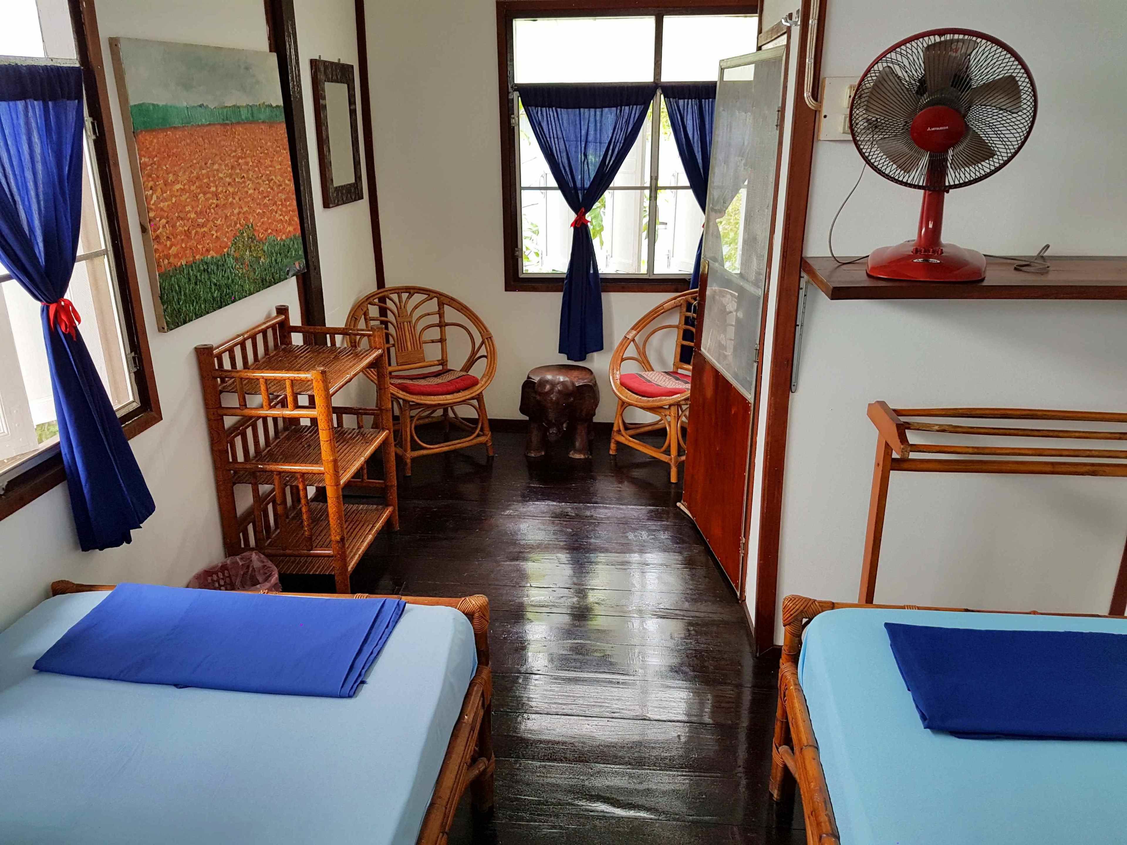 Une chambre avec deux lits, une table, des chaises, un ventilateur et une fenêtre avec des rideaux.