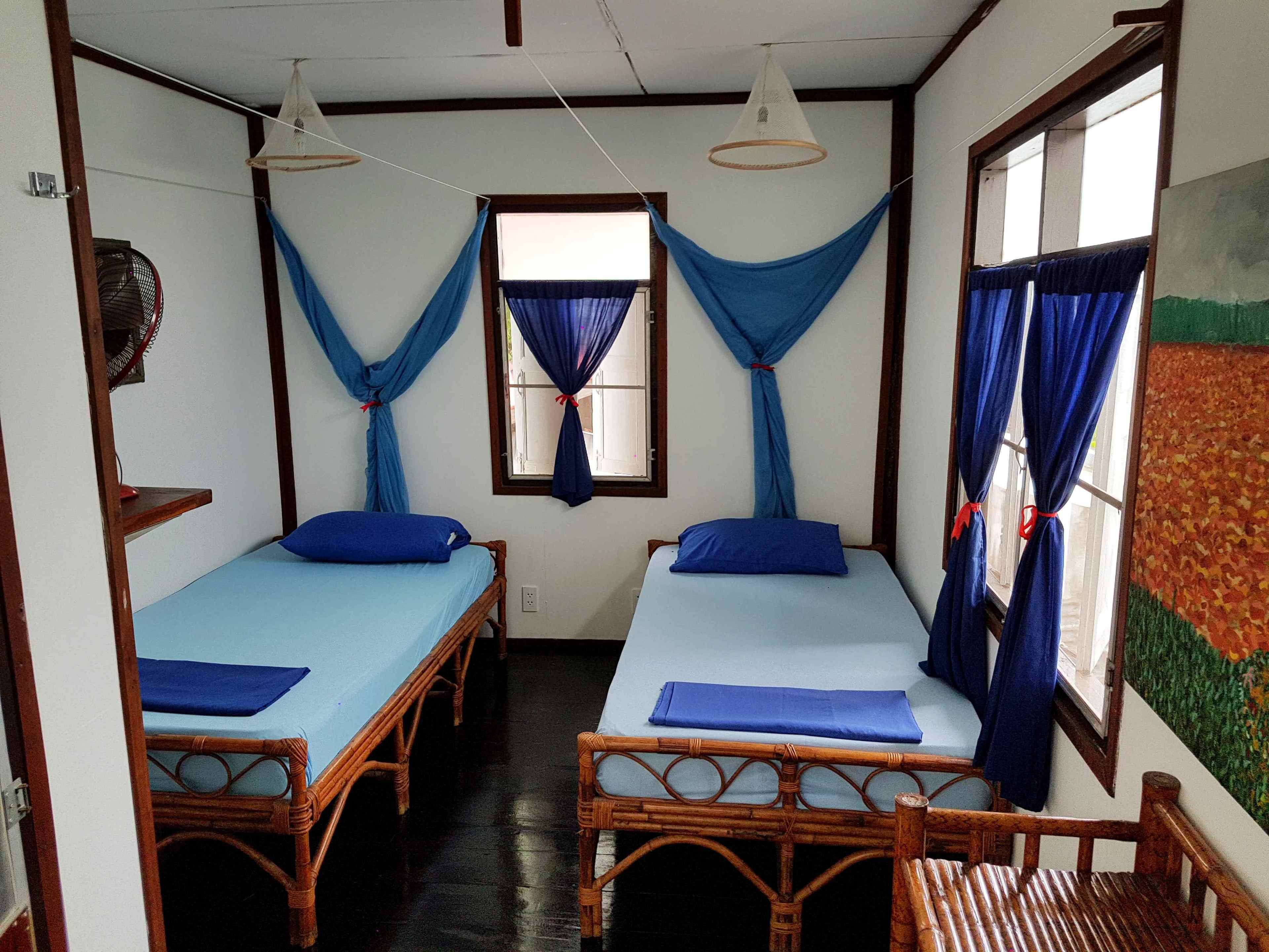 A room with two wooden beds, each with a blue mattress and pillow, and a window with blue curtains. There is a small table with a blue tablecloth and a chair.