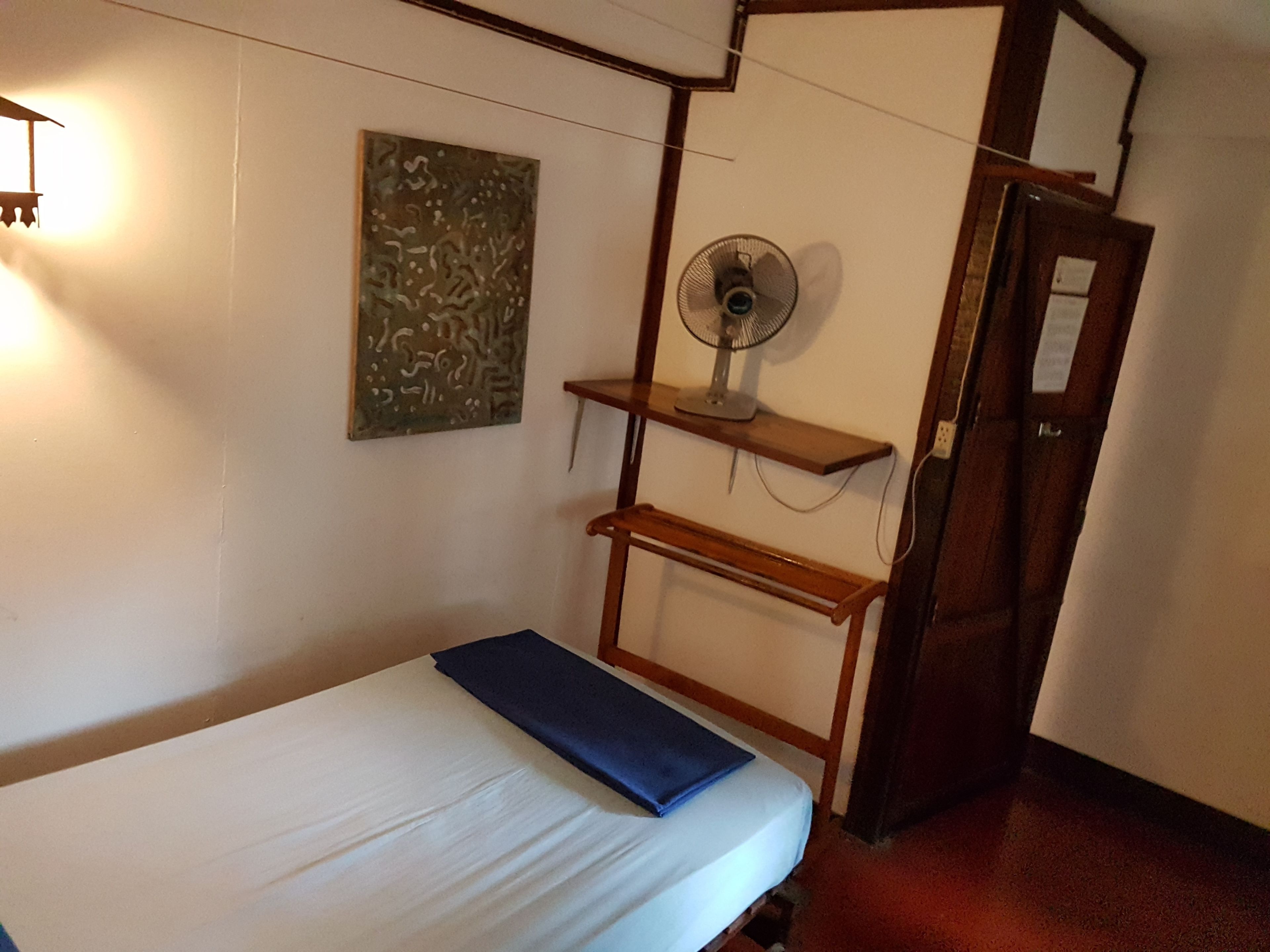 A single bed with a blue blanket, a wall-mounted fan, a shelf with a picture, and a door to another room.