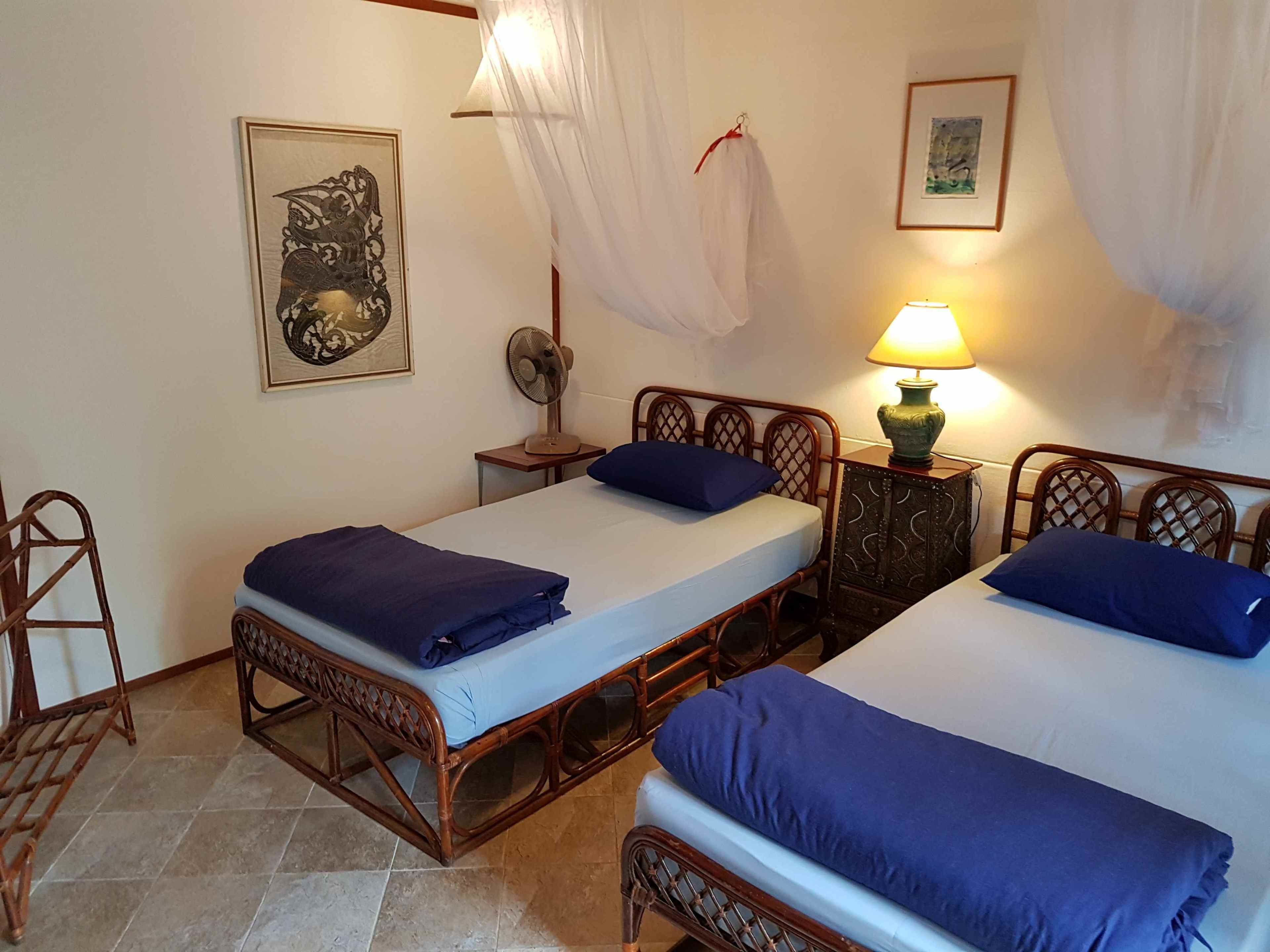 Una habitación con dos camas de madera, cada una con colchón y almohada azules, un ventilador de pie, una mesita de noche con lámpara y un cuadro en la pared.
