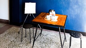 Quarto com parede azul escura, uma mesa de madeira com cafeteira, xícaras e um quadro branco. Há dois taburetes pretos e um pendente.