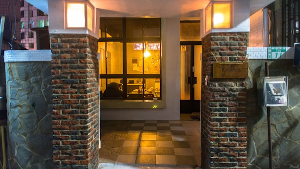 Entrada de hotel com paredes de pedra e tijolo, chão xadrez e uma janela com iluminação interna.