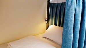 Uma cama de solteiro com roupa de cama branca, uma cortina escura e um interruptor na parede.