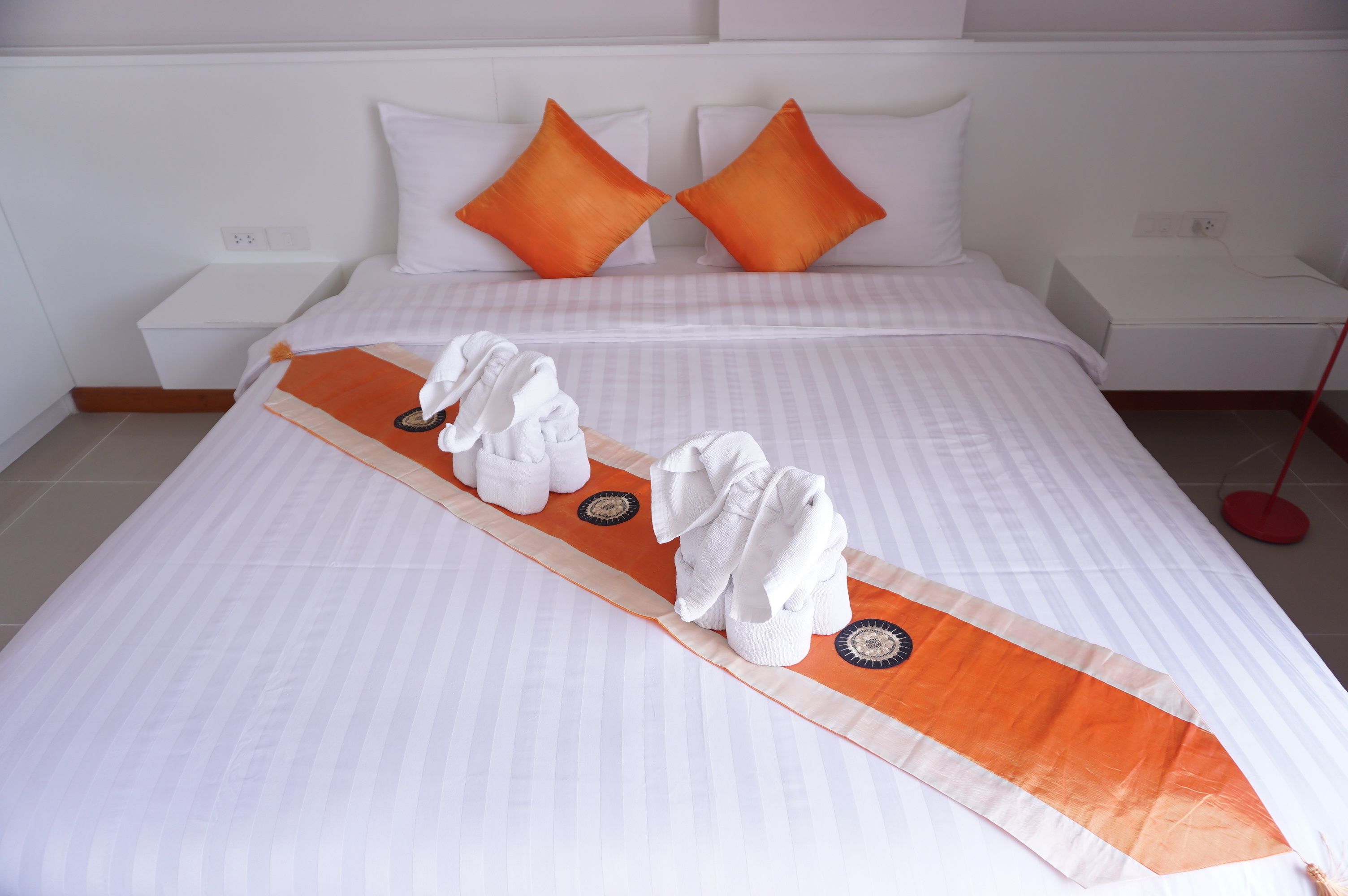A neatly made bed with white linens and an orange runner, two orange pillows, and a white bedside table with a red floor lamp.