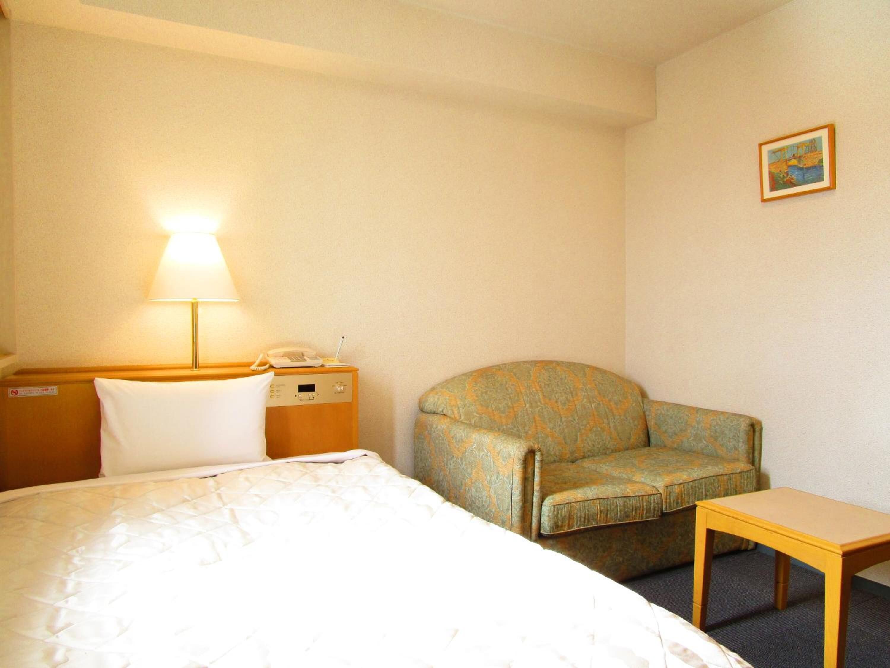 A hotel room with a bed, a chair, a small table, a lamp, and a phone on a bedside table.