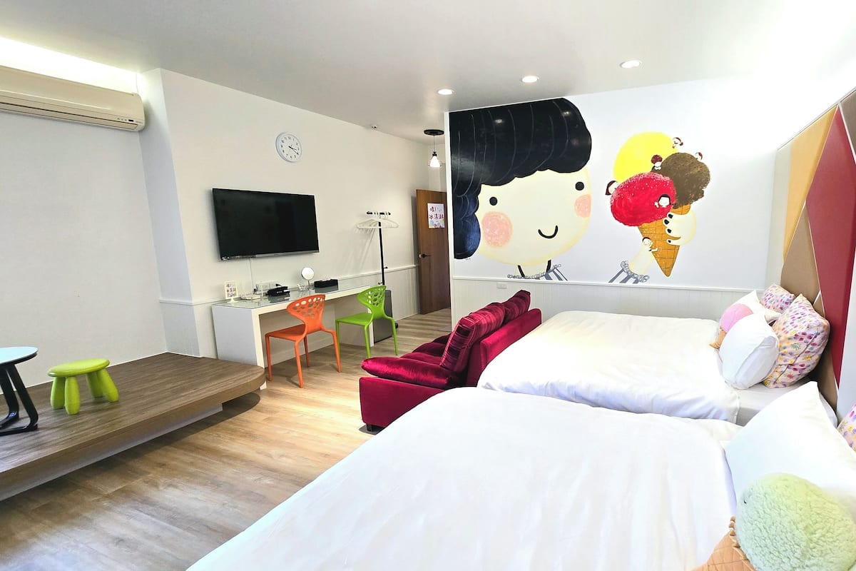 Habitación de hotel con dos camas, un escritorio con silla, un televisor y un mural en la pared de una niña con un helado.