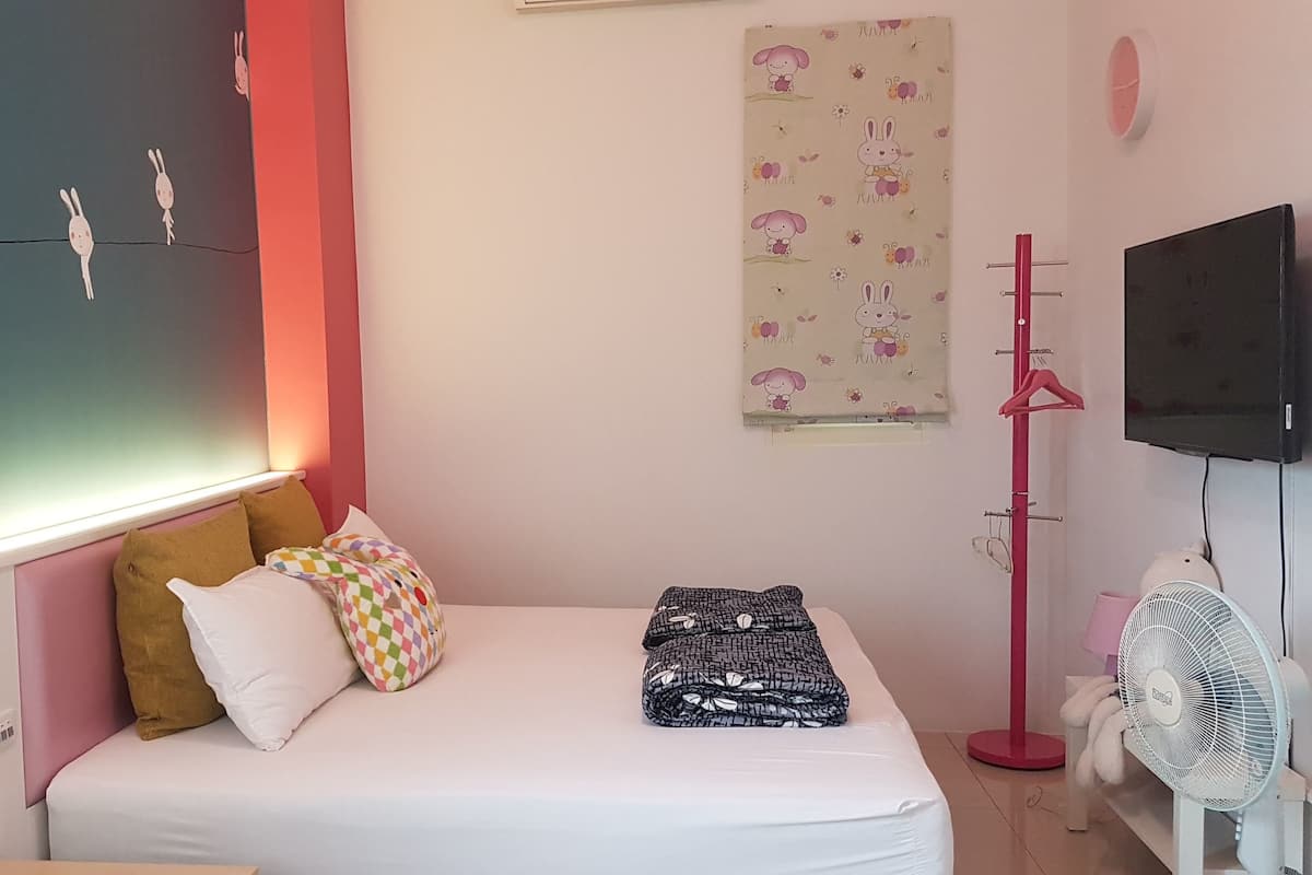 Un dormitorio con cama, almohadas, una pared con diseño, televisor y ventilador.