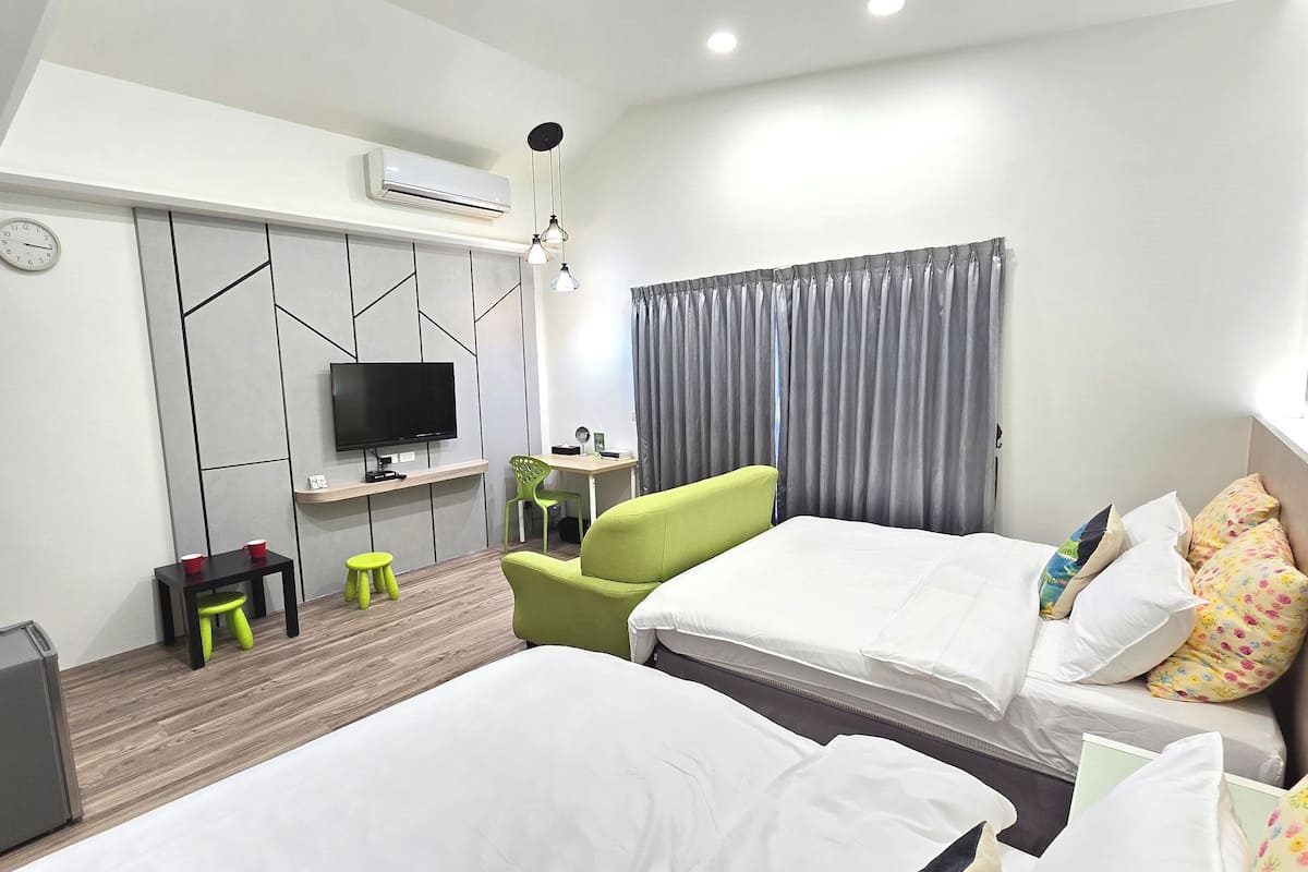 Habitación de hotel con dos camas, un escritorio con televisor de pantalla plana, una pequeña zona de estar con taburetes y una ventana con cortinas.