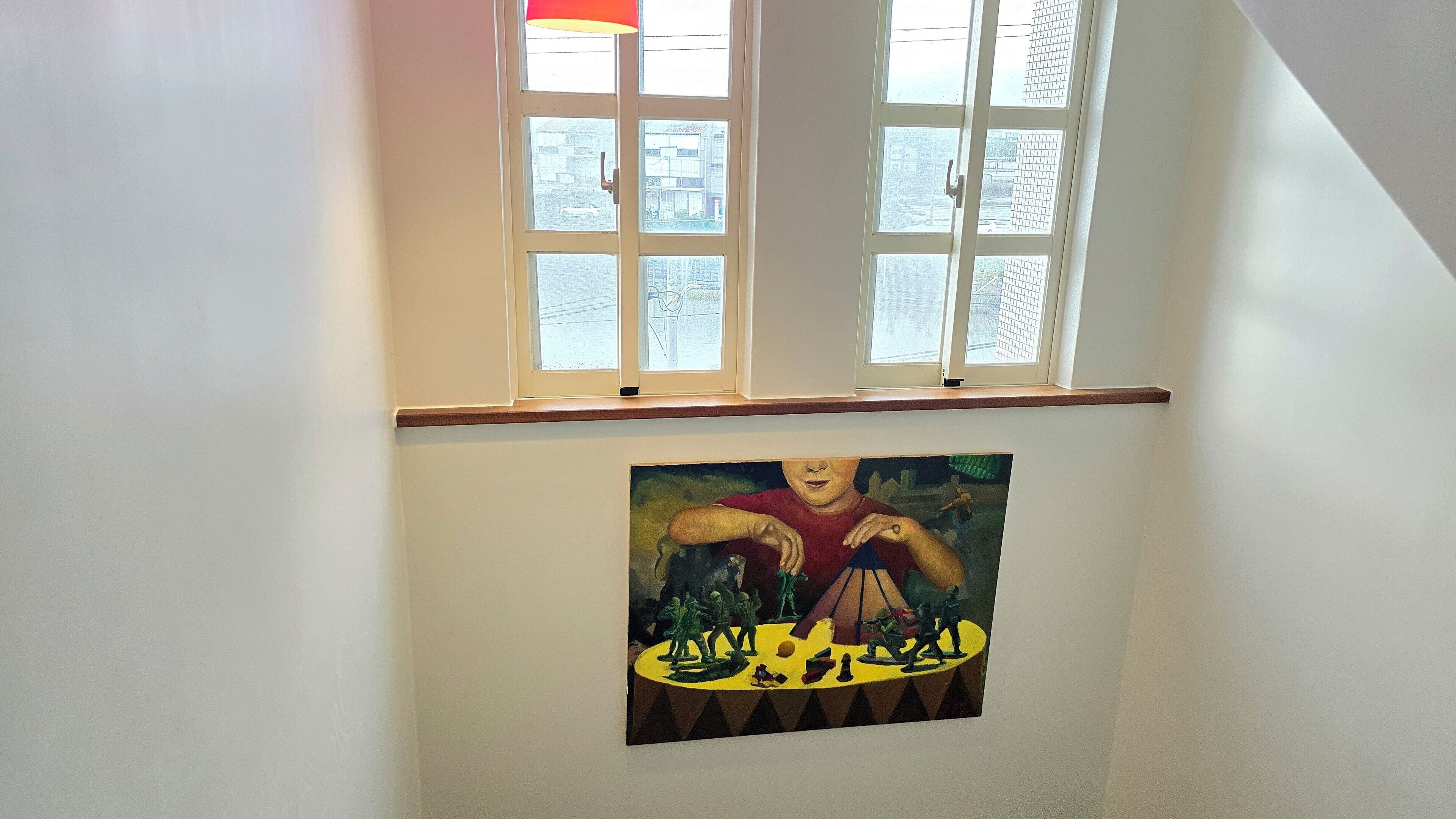 Ein Zimmer mit zwei Fenstern, einem Bild an der Wand und einem Holzbrett über den Fenstern.