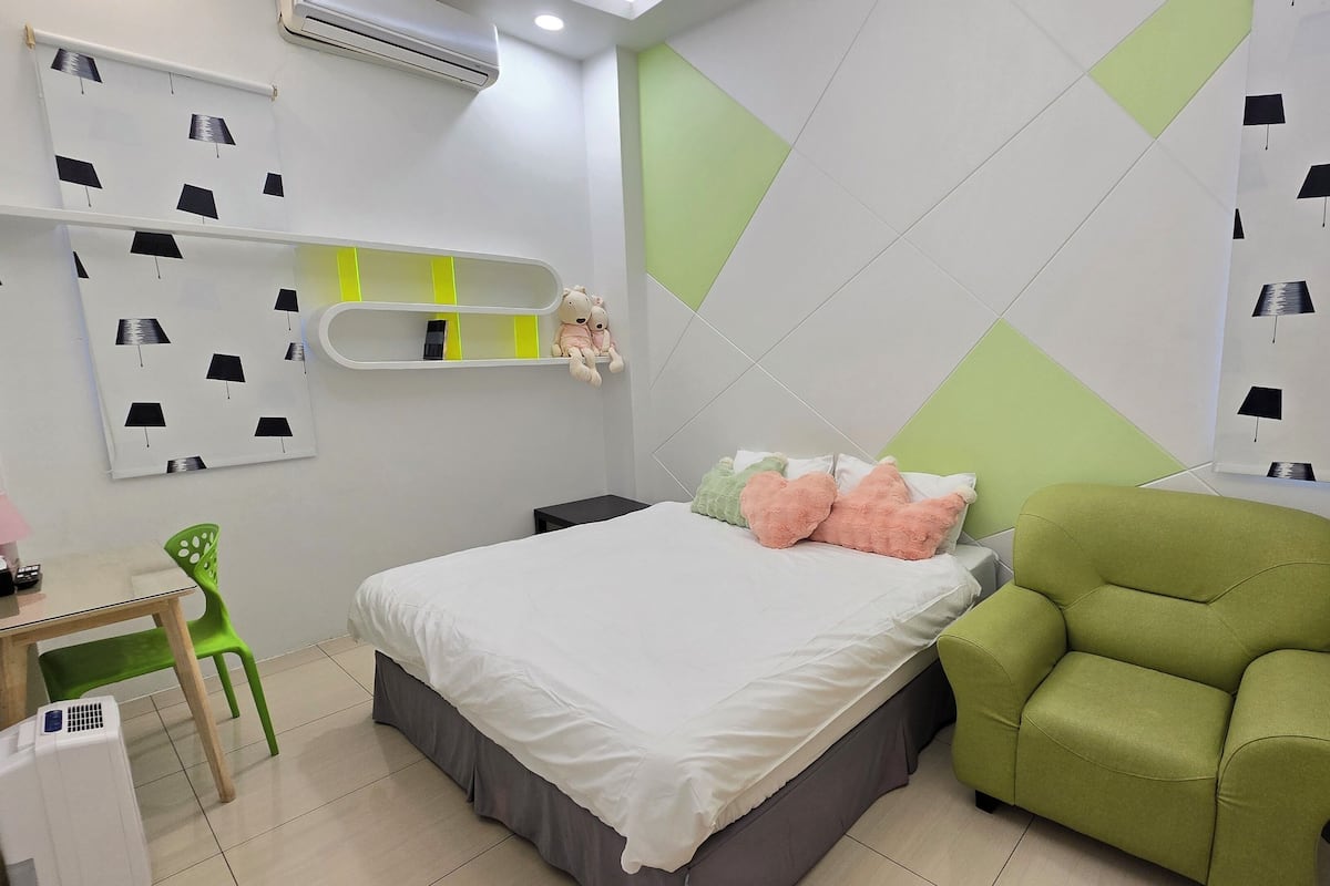 Un dormitorio con una cama, un sillón verde, una mesita y un estante en la pared con un osito de peluche.