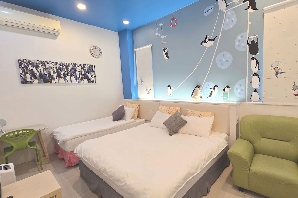 Una habitación con dos camas, una silla verde, un reloj de pared y una pared decorada con pingüinos.