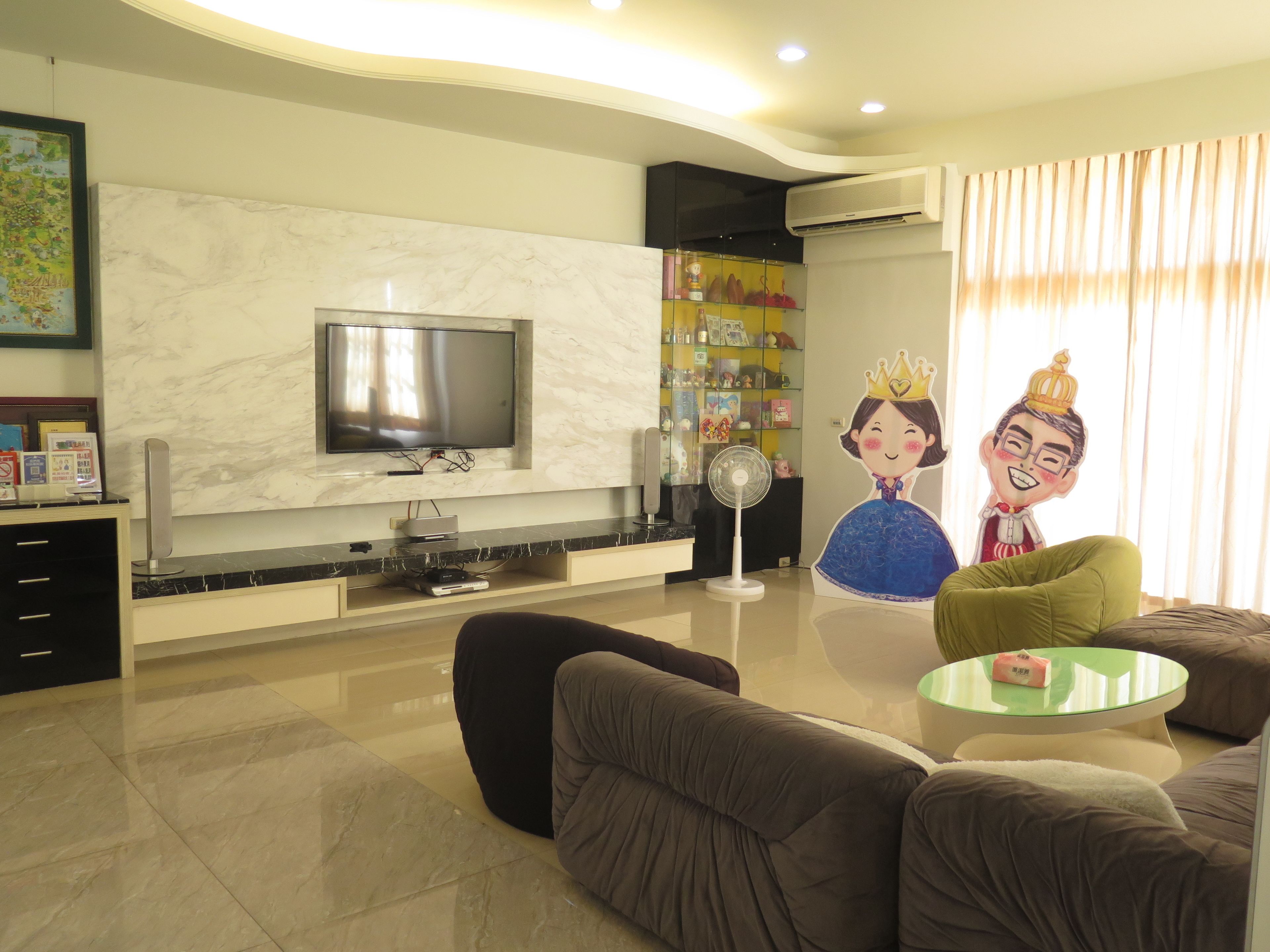 A modern living room with a flat-screen TV, comfortable seating, a glass coffee table, and decorative elements including a wall-mounted fan and a large poster of cartoon characters.