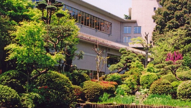 Um edifício com arquitetura moderna, rodeado por vegetação exuberante e um lago espelhado.