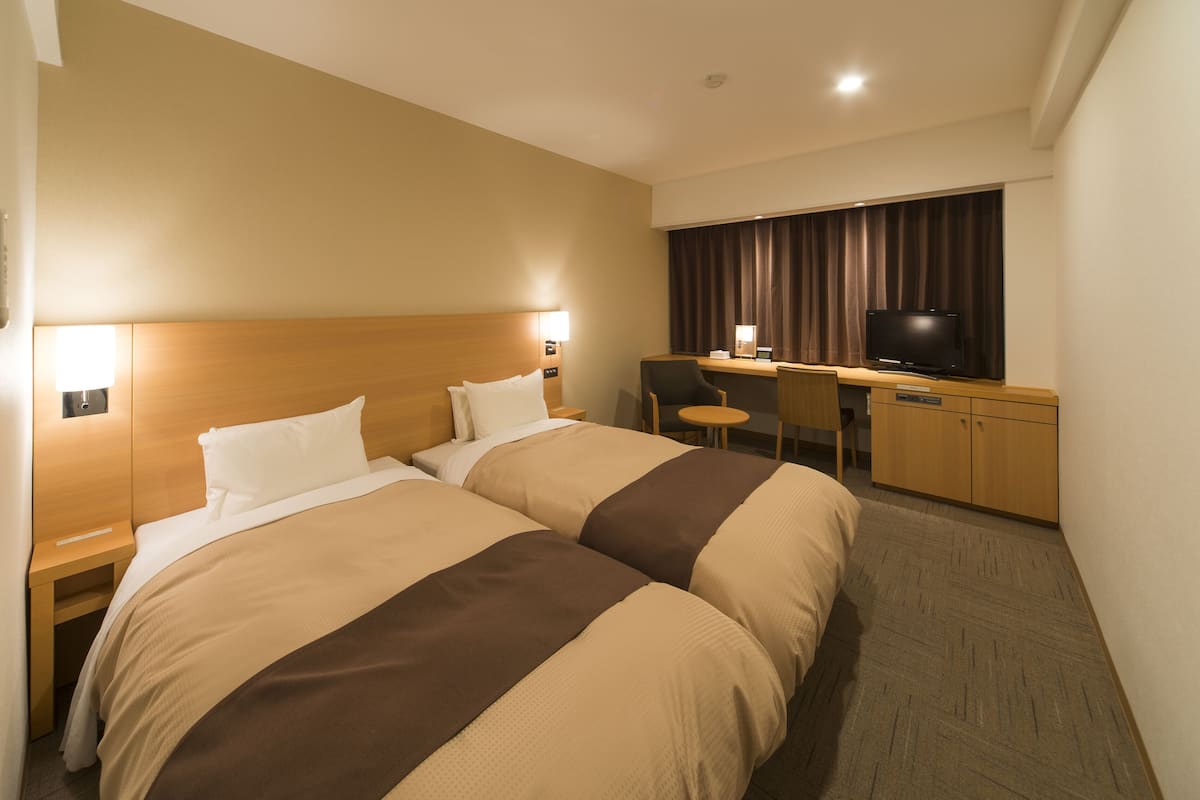 Ett hotellrum med två sängar, ett skrivbord med en stol, en tv och ett fönster med gardiner.