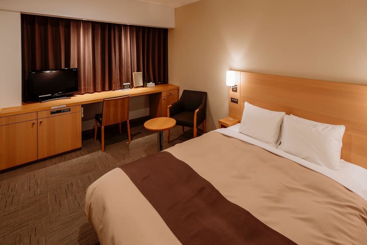 Ett hotellrum med en säng, ett skrivbord med en stol, en tv och en lampa.