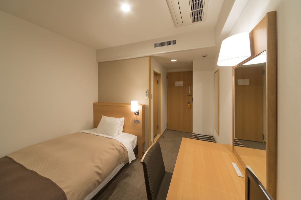 Ett hotellrum med en säng, ett skrivbord i trä, en stol, en spegel och en dörr som leder till ett annat rum.