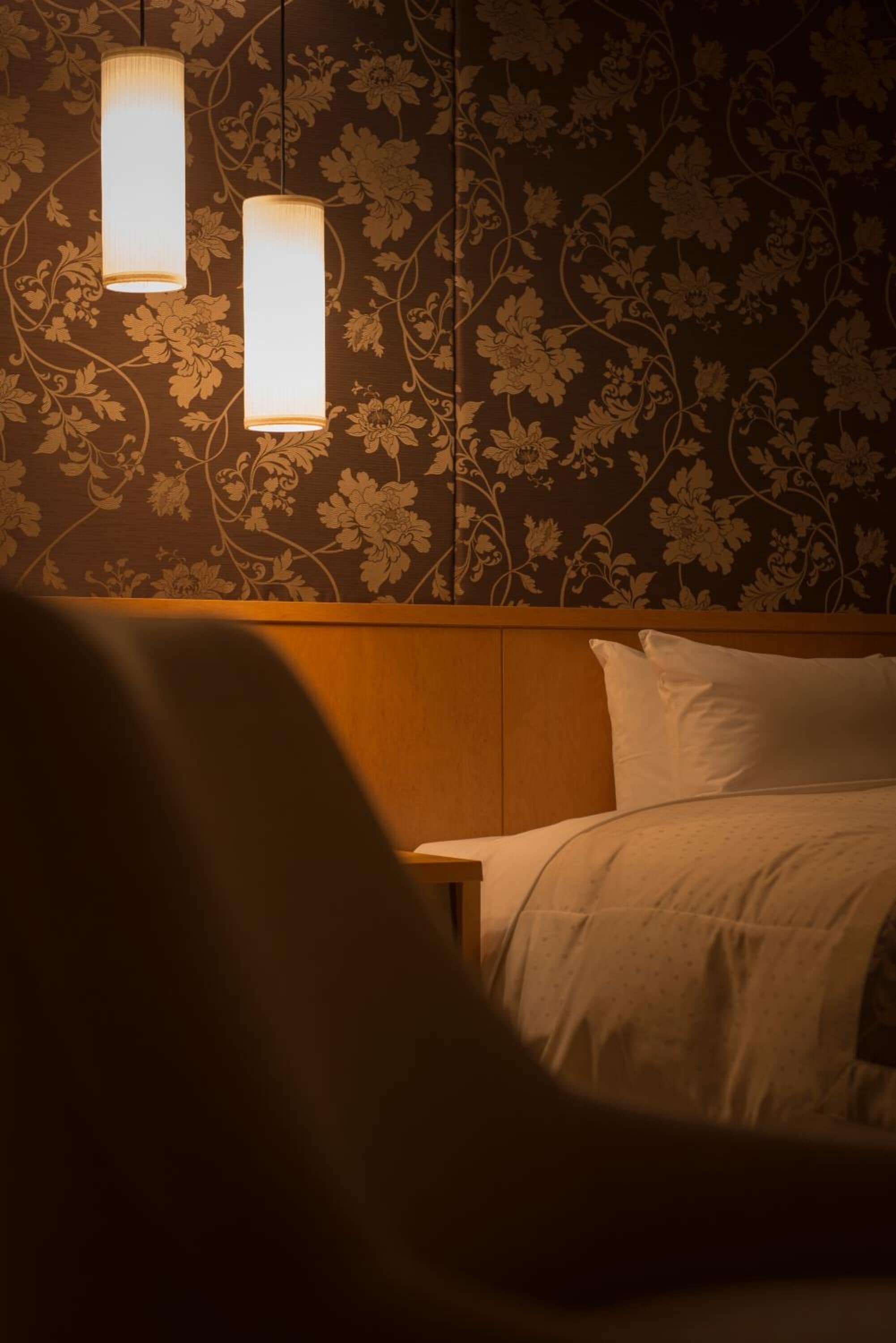 Een hotelkamer met bloemenbehang, een houten hoofdeinde en twee hangende hanglampen.