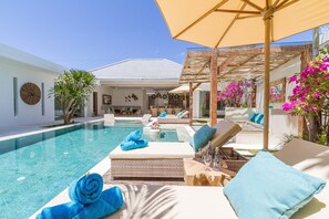 Zona de piscina con sillones de descanso, un pérgola y vista a un edificio moderno.