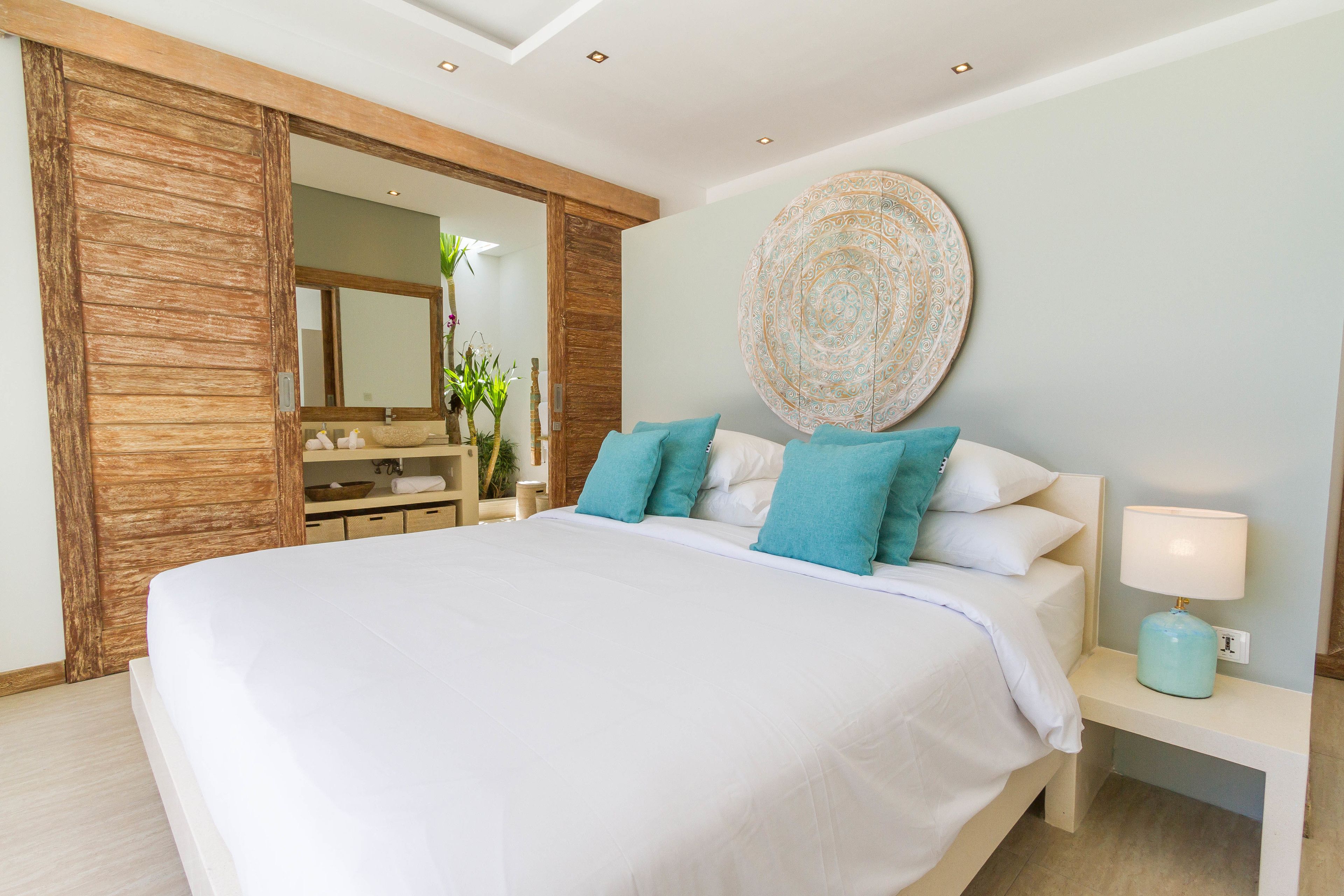 Een groot bed met witte beddengoed en teal kussens, een houten garderobe, een rond decoratief object aan de muur en een nachtkastje met een lamp.