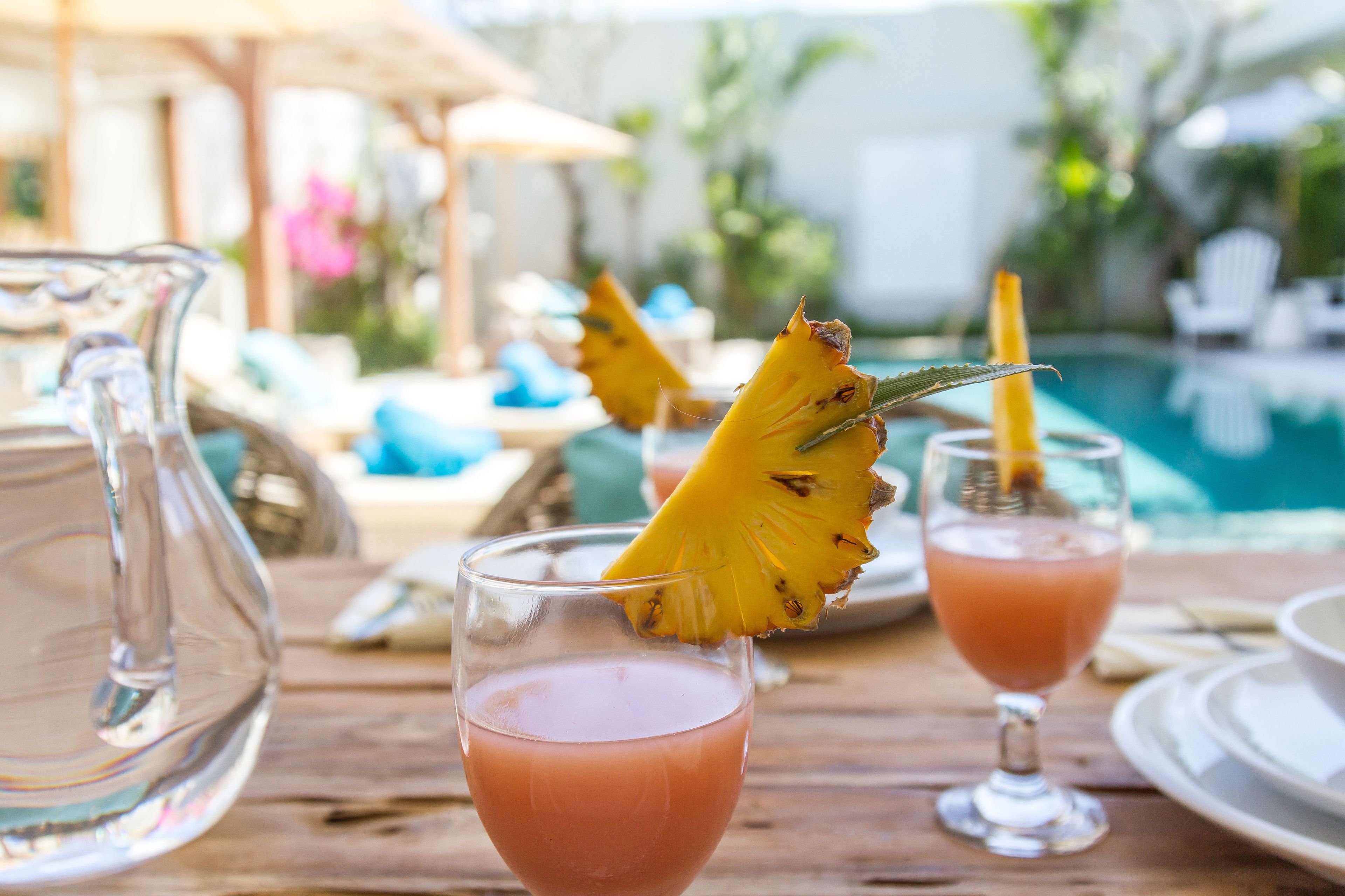 Ein Holztisch mit zwei Gläsern rosafarbenem Getränk, jedes mit einer Ananas Scheibe verziert. Auf dem Tisch steht auch eine Karaffe.