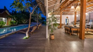 A wooden deck with a pool, outdoor furniture, and a dining area with a ceiling fan and pendant lights. - Villa Nahema (Seminyak)