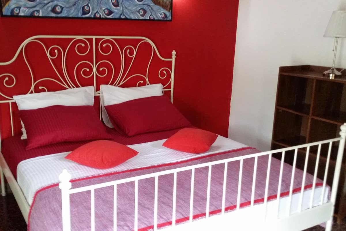 A bed with a white metal frame and red bedding, a red wall, and a painting above the bed.