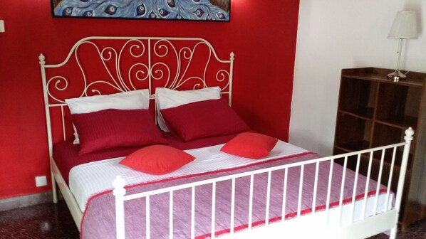 A bed with a white metal frame and red bedding, a red wall, and a painting above the bed. - Village Villa Home Stay (Kandy)
