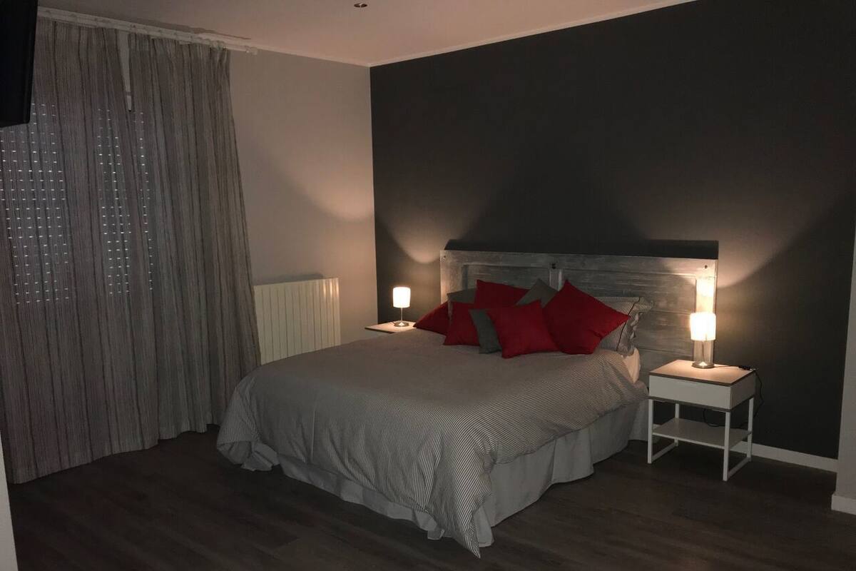 A bedroom with a bed, two bedside tables with lamps, a window with curtains, and a dark wall.