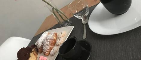 A table with a plate of pastries, a black cup, a glass, and a vase with a red flower.