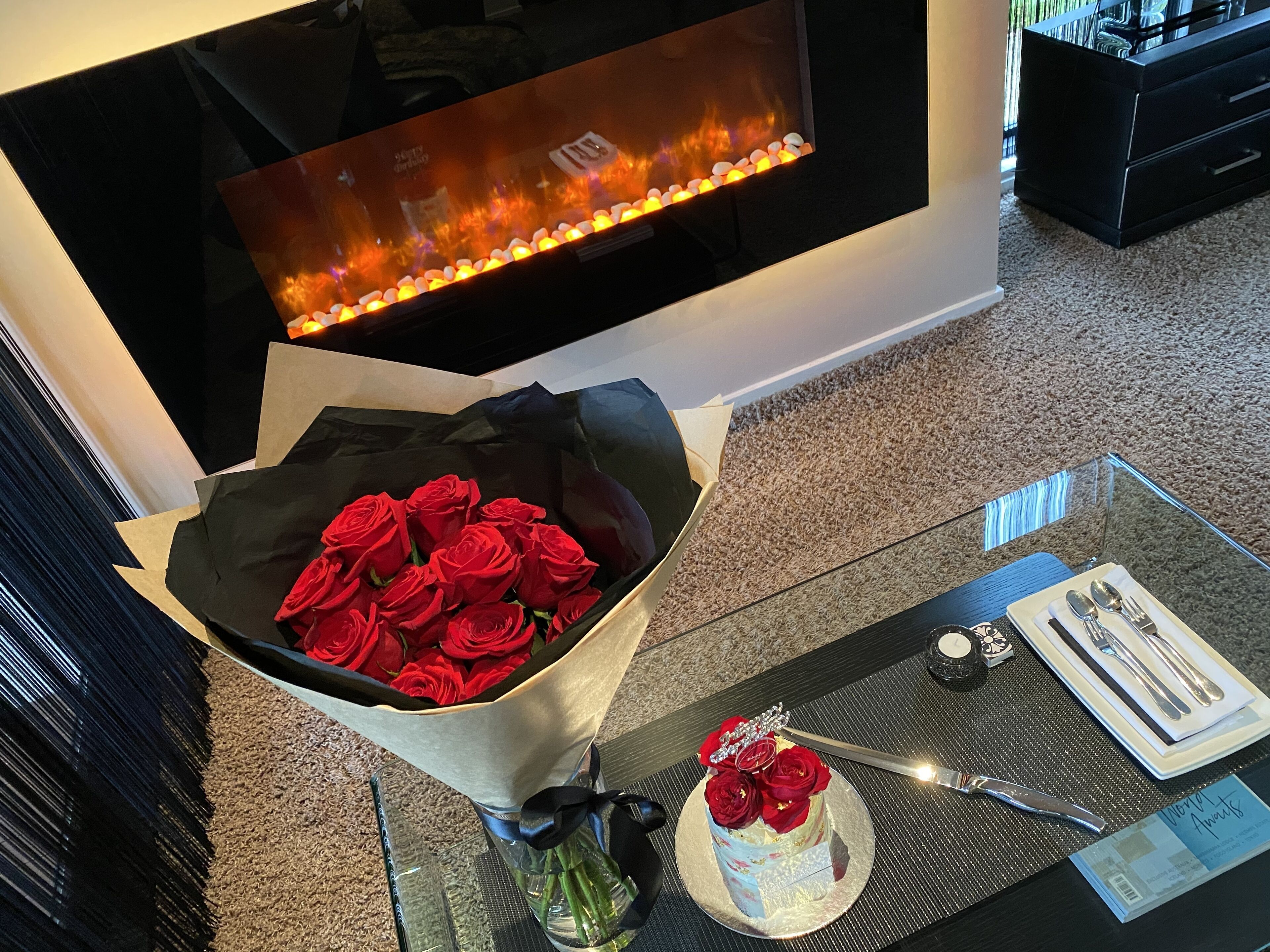 A room with a fireplace, a bouquet of red roses, a cake with roses, and a table set with cutlery.