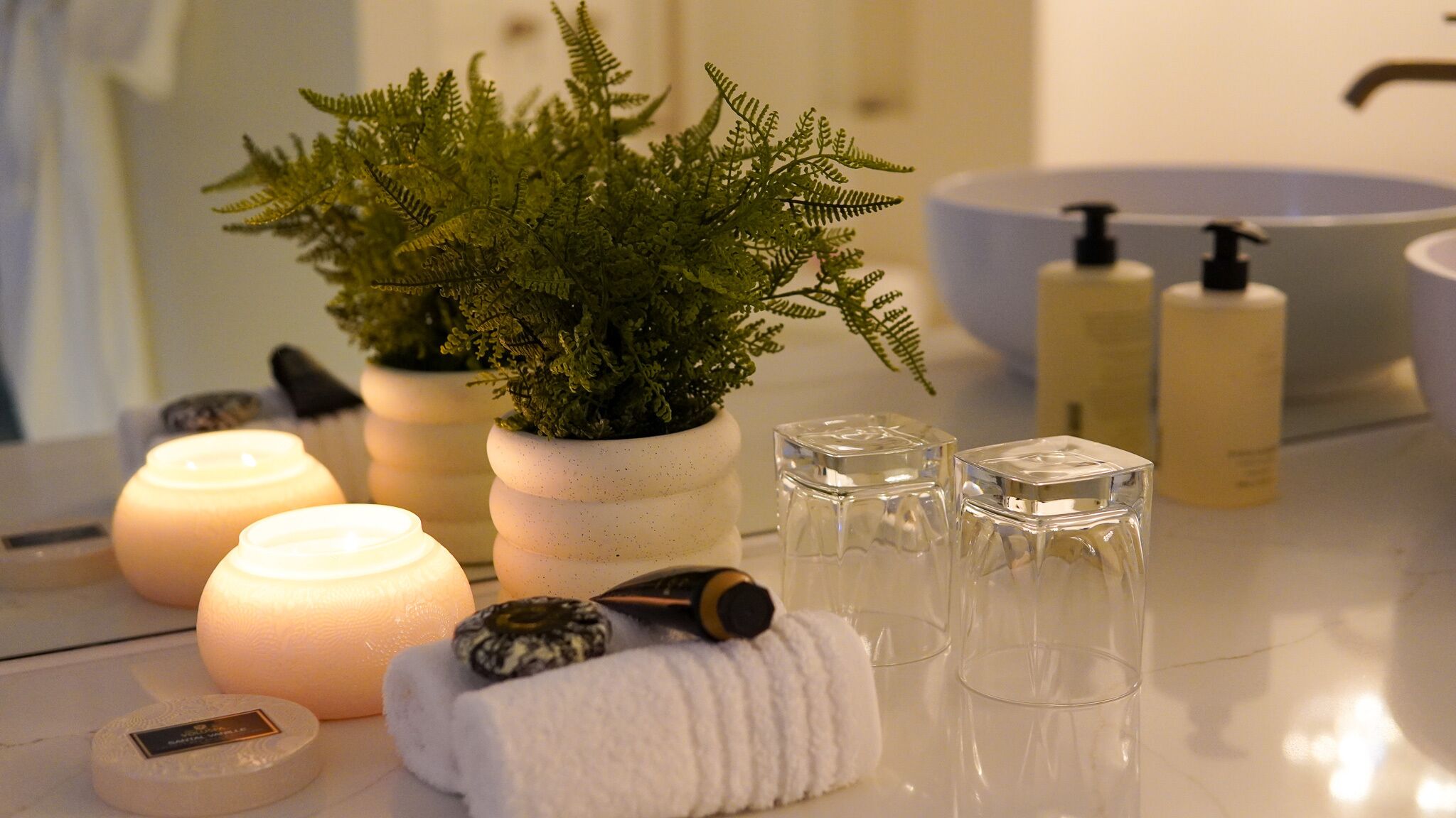 Uma casa de banho com uma pia branca, duas embalagens de sabonete líquido, uma planta em vaso, velas e uma toalha.