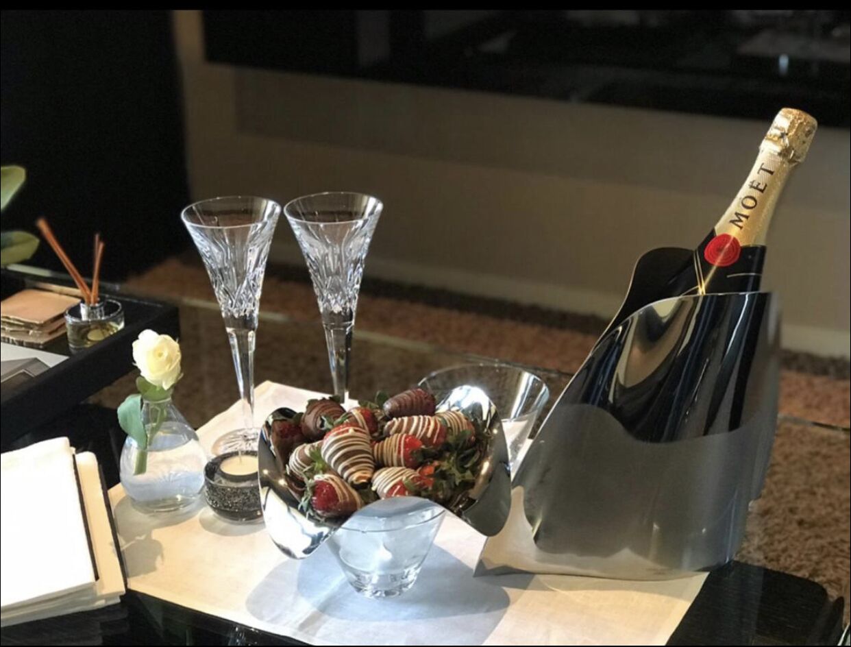 A table with a bottle of Moët champagne, two crystal glasses, a bowl of chocolate-covered strawberries, and a small vase with a white rose.