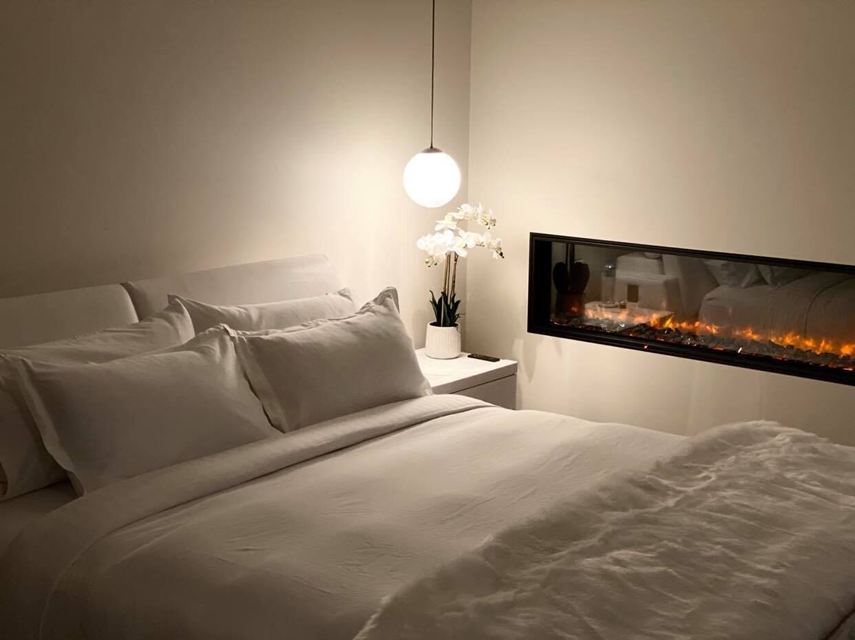 Un dormitorio moderno con una cama grande, una mesita de noche con lámpara y un televisor de pared mostrando un fuego.