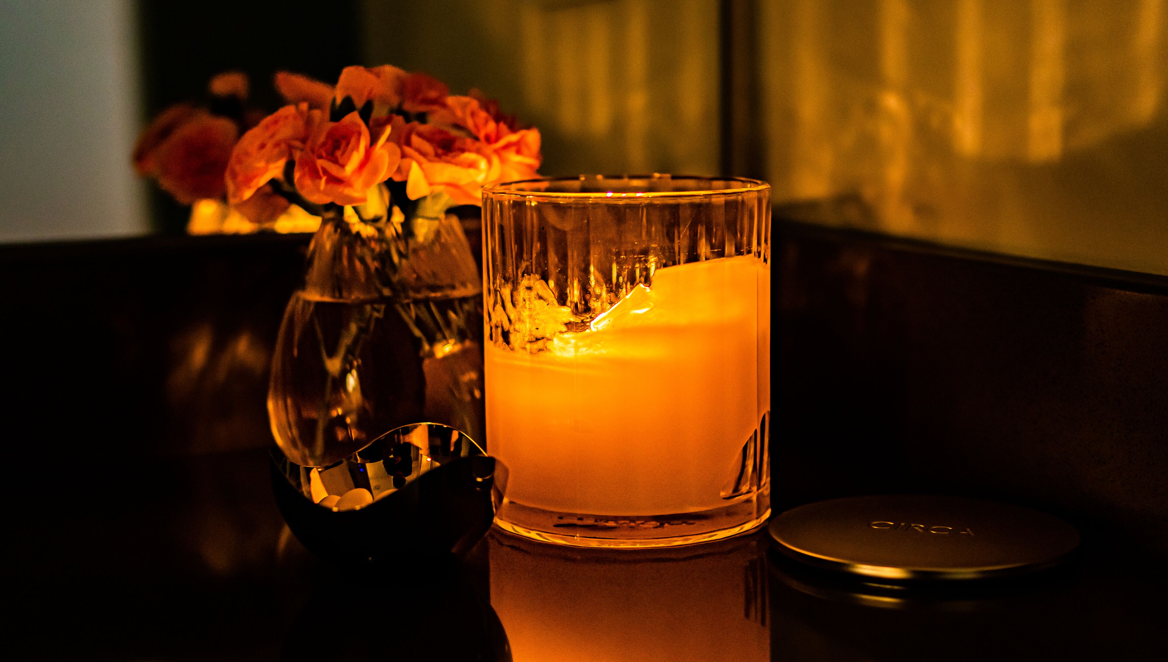 A candle in a glass holder on a table, with a vase of flowers in the background.