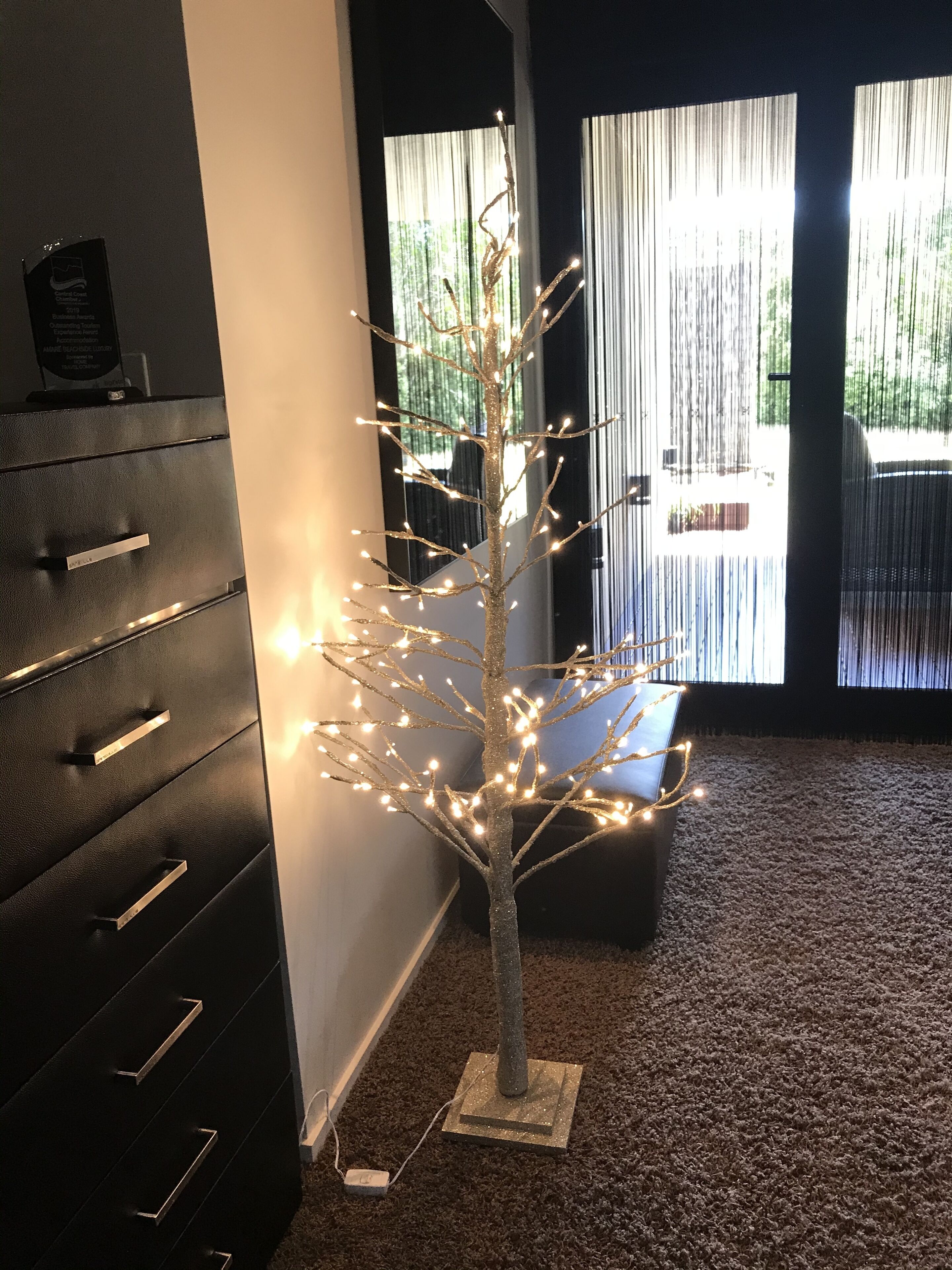 Une pièce décorée d’un arbre illuminé, avec un meuble de rangement noir et une vue dégagée sur l’extérieur grâce à de grandes fenêtres.