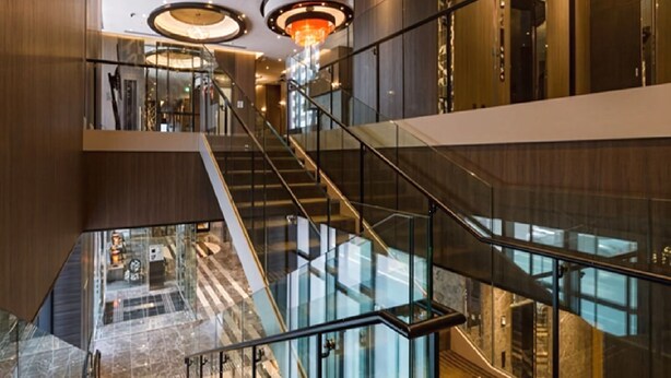 Un interior moderno con una escalera de cristal que conecta varios pisos, barandillas metálicas y un candelabro central.