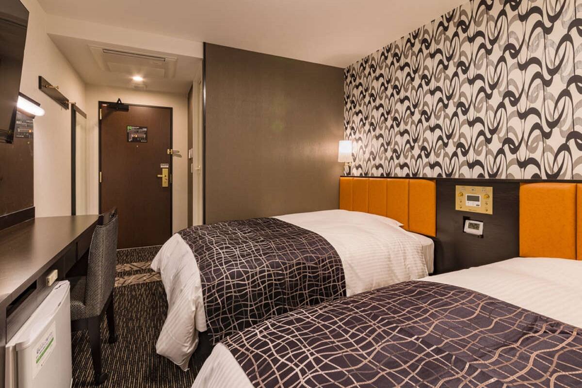 A hotel room with two beds, a desk, and a chair. There is a door with a sign, a patterned wall, and a control panel on the headboard.
