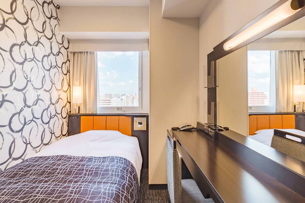 A hotel room with a bed, a desk, a mirror, and a window with a city view.