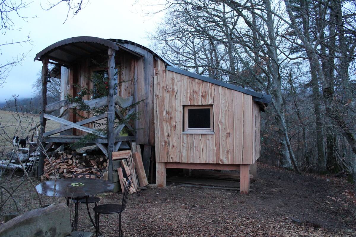 Una cabaña de madera con techo inclinado, una ventana pequeña y una puerta. Hay una pila de leña y una mesa de metal con sillas afuera.