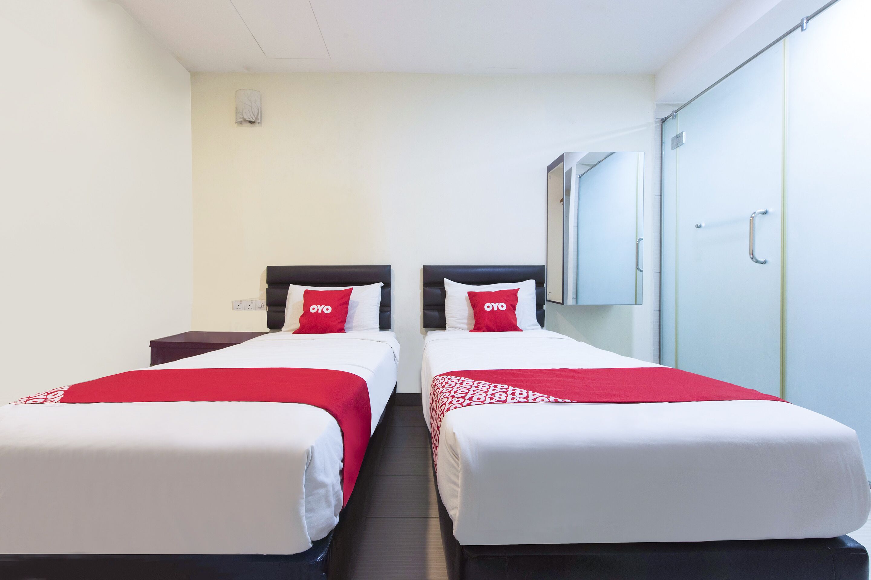 A hotel room with two beds, each with a red and white bedspread and pillows, a small bedside table, and a mirror on the wall.