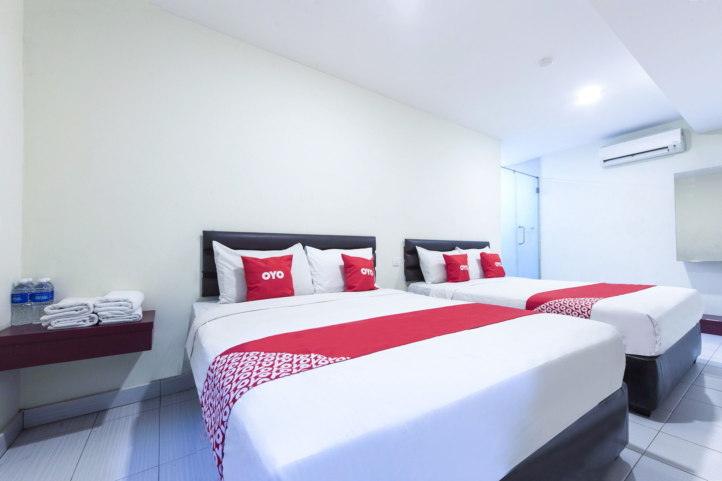 Quarto de hotel com duas camas, cada uma com roupa de cama branca e almofadas vermelhas, um cobertor estampado em vermelho e branco, uma pequena prateleira com toalhas e produtos de higiene pessoal, e um aparelho de ar condicionado na parede.