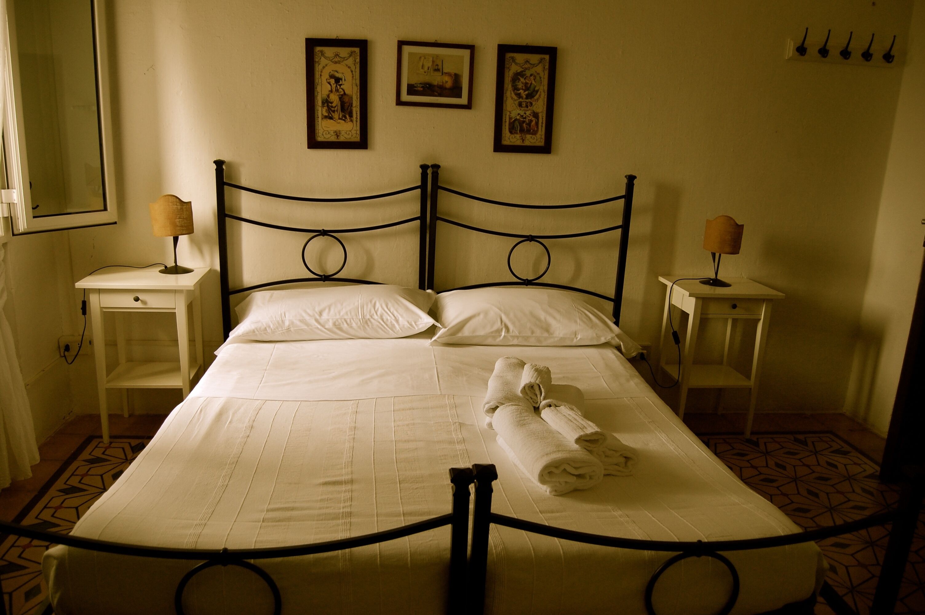 Una cama bien hecha con ropa blanca y una toalla doblada, dos mesitas de noche con lámparas, y una pared con tres cuadros enmarcados.