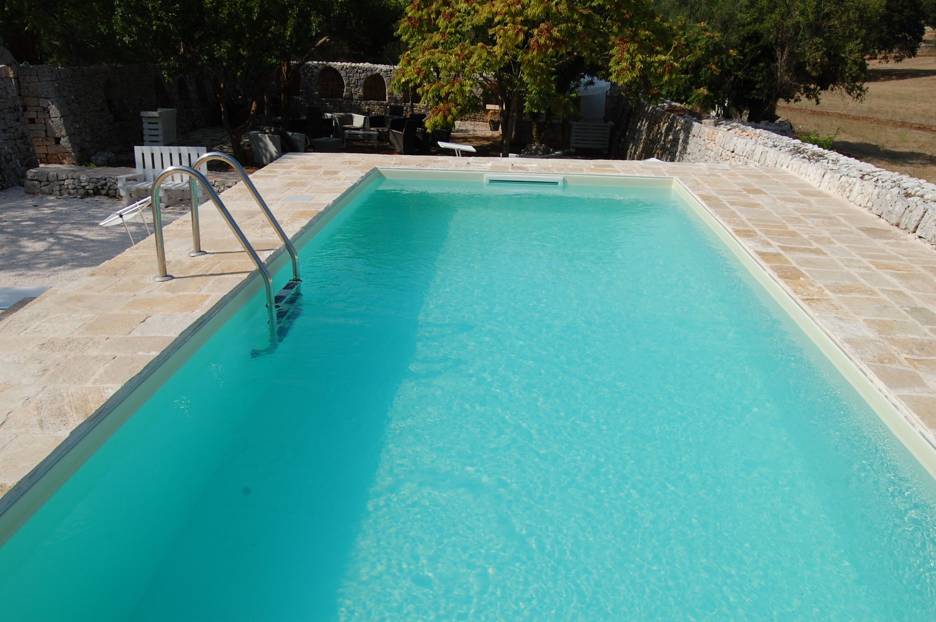 A rectangular swimming pool with clear blue water, surrounded by stone tiles and a stone wall.