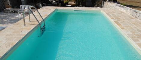 A rectangular swimming pool with clear blue water, surrounded by stone tiles and a stone wall.