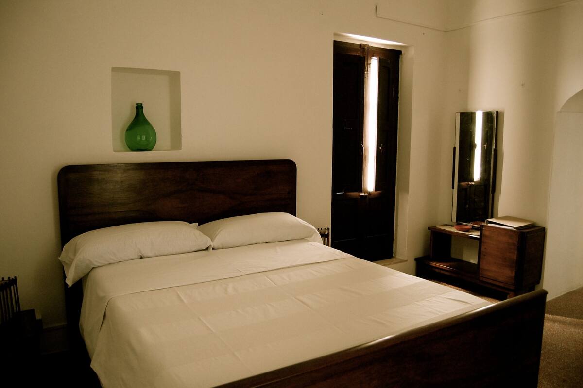 A neatly made bed with white linens, a wooden headboard, a green vase on the wall, and a mirror on a wooden dresser.