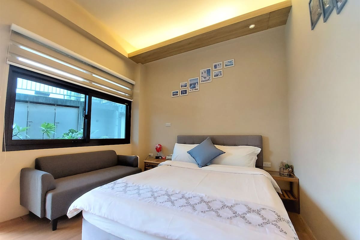Un dormitorio moderno con una cama grande, un sofá, una ventana con persianas y una pared decorada con cuadros.