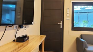 Habitación con televisión de pantalla plana montada en la pared, una mesa de madera con un control remoto, una puerta oscura y una ventana con vistas a plantas afuera.