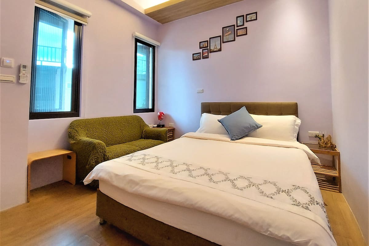 Un dormitorio con una cama, un sillón verde, una mesita de noche y una pared con marcos.