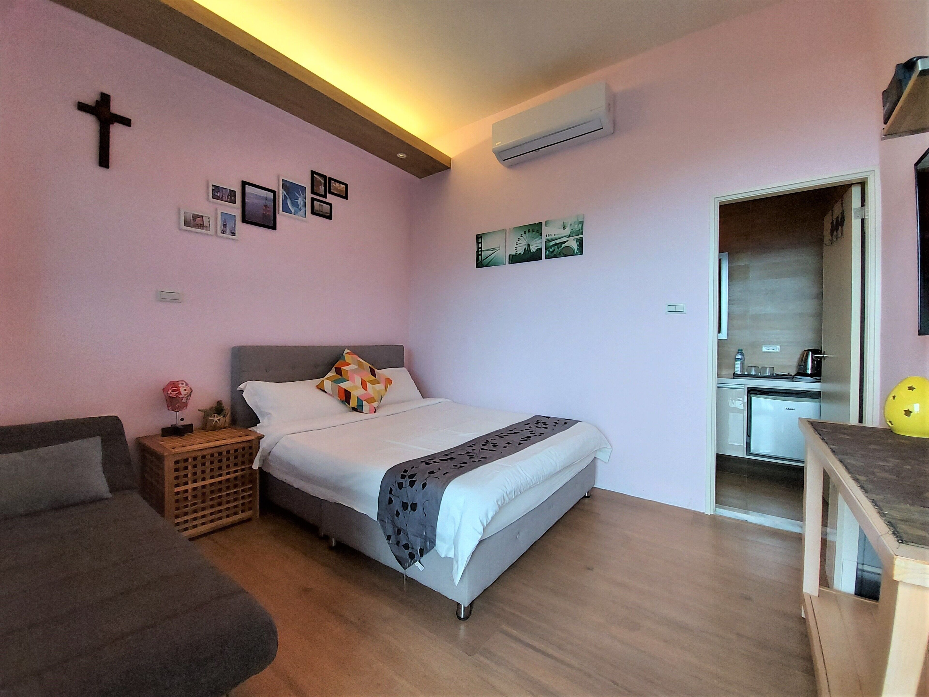 A bedroom with a bed, a sofa, a small table, a wall-mounted air conditioner, and a kitchen area visible through an open door.